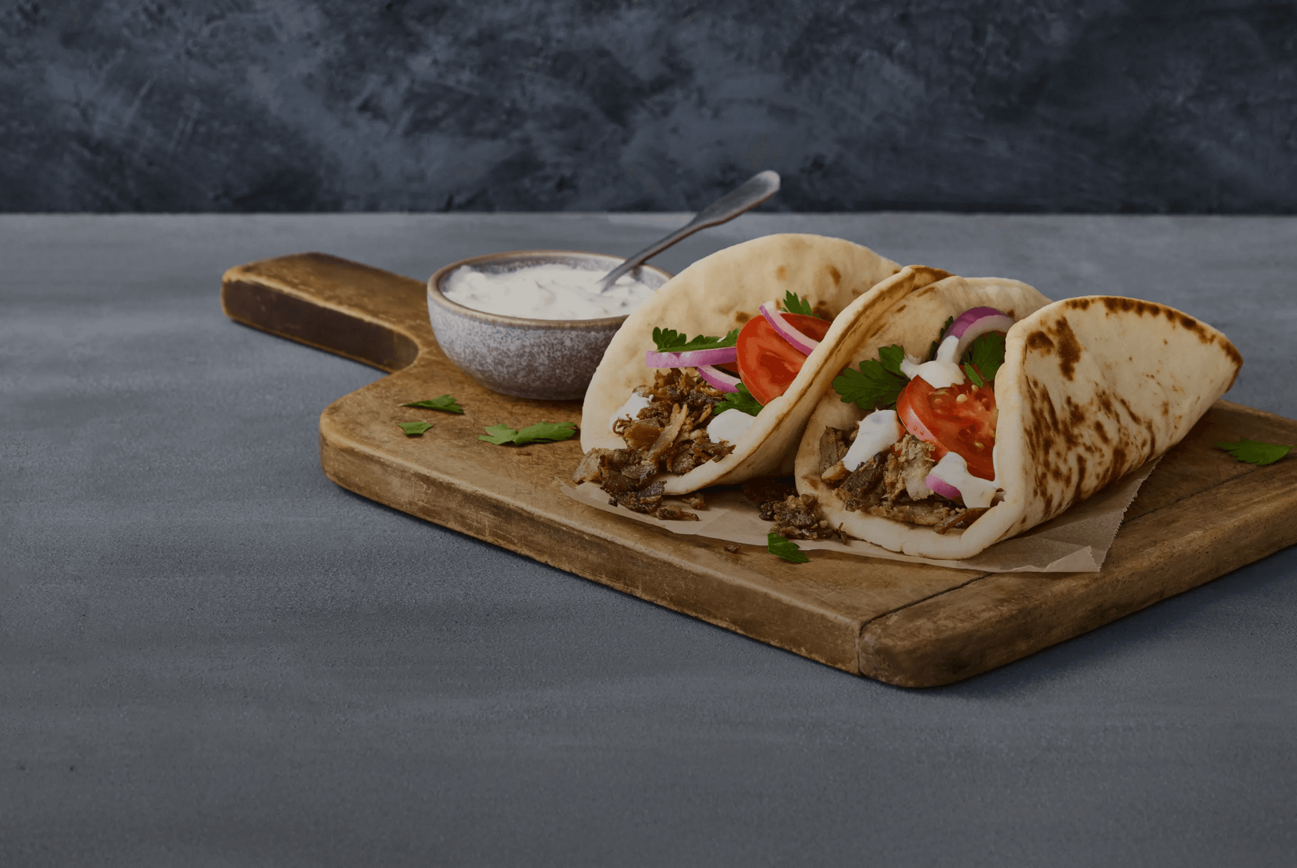 Two pita sandwiches filled with meat, tomato slices, onion, and herbs are placed on a wooden board, alongside a bowl of white sauce with a spoon on a gray surface.