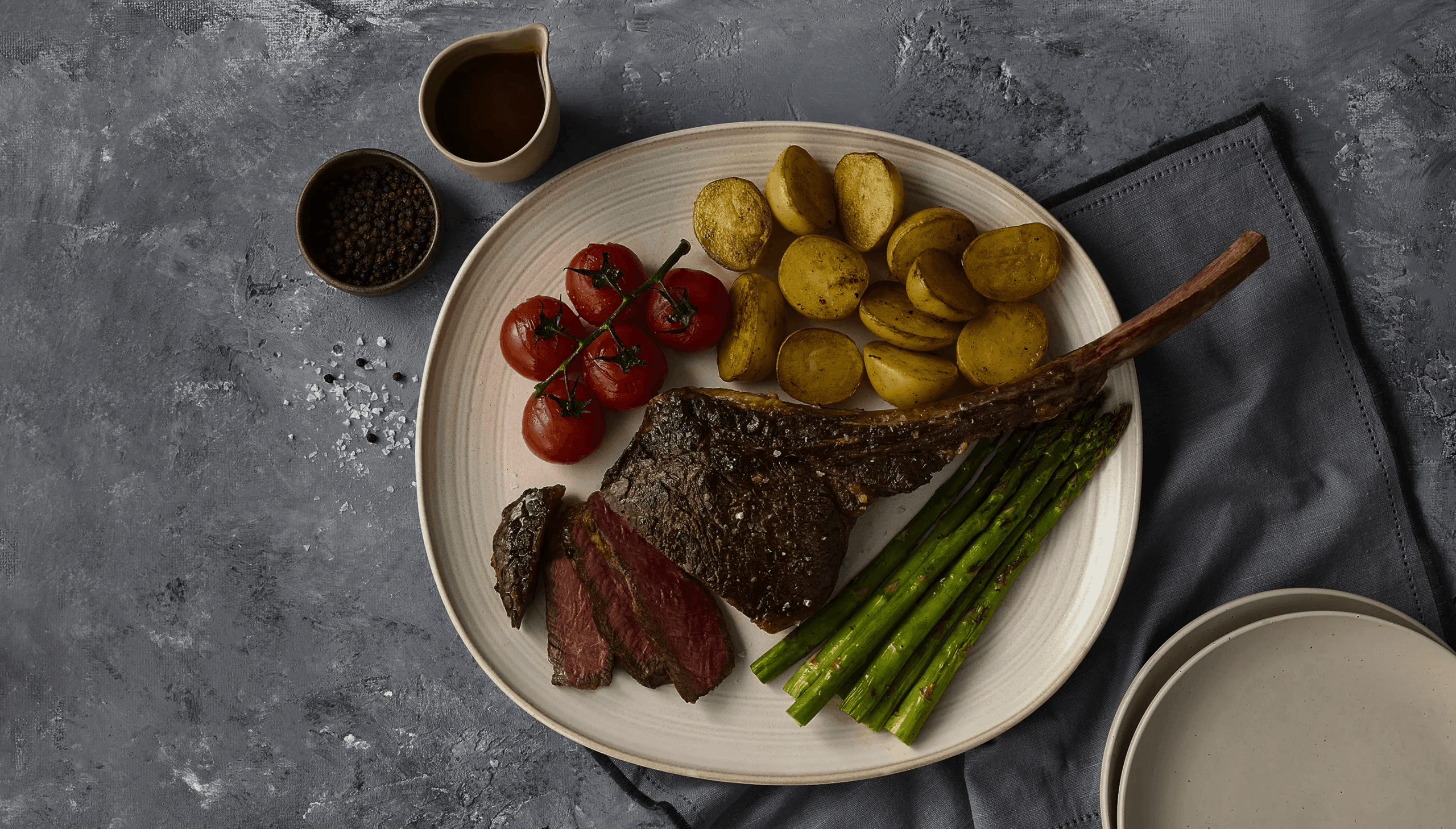 Tomahawk steak dinner: steak, asparagus, potatoes, tomatoes, peppercorns, and sauce on a plate.