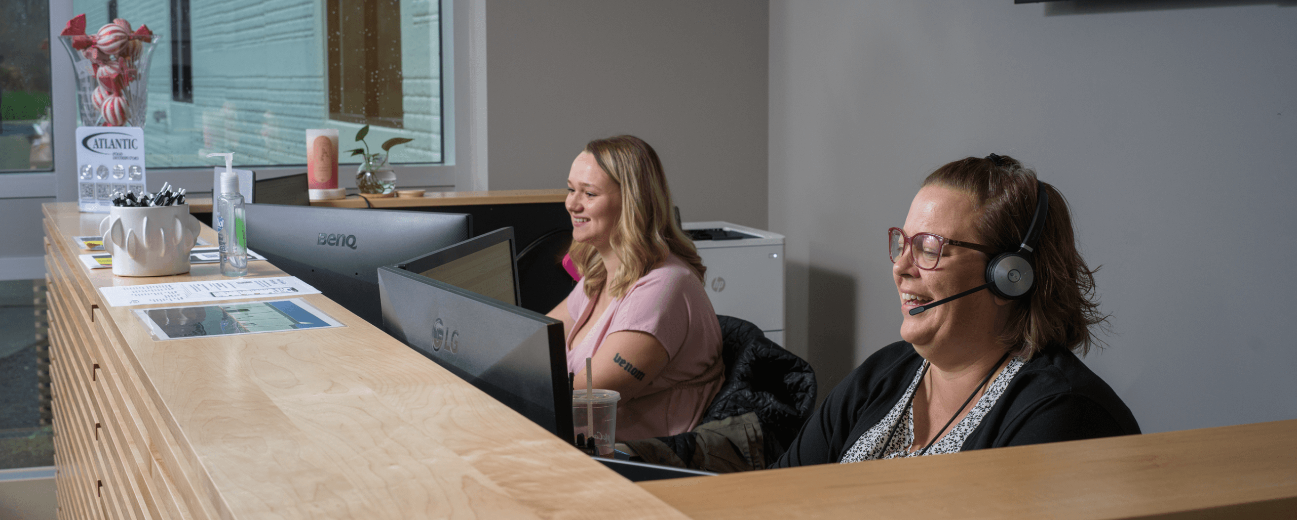 Two friendly receptionists at a modern office desk, one wearing a headset.