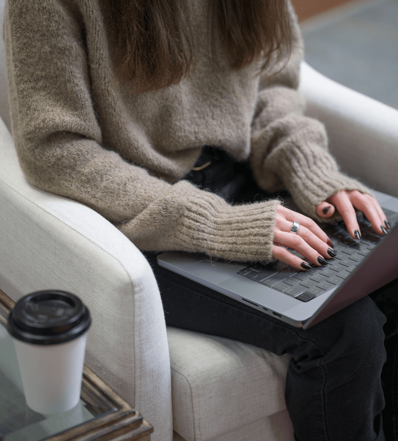Person typing on a laptop while seated in a cozy armchair, wearing a beige sweater, with a coffee cup nearby.