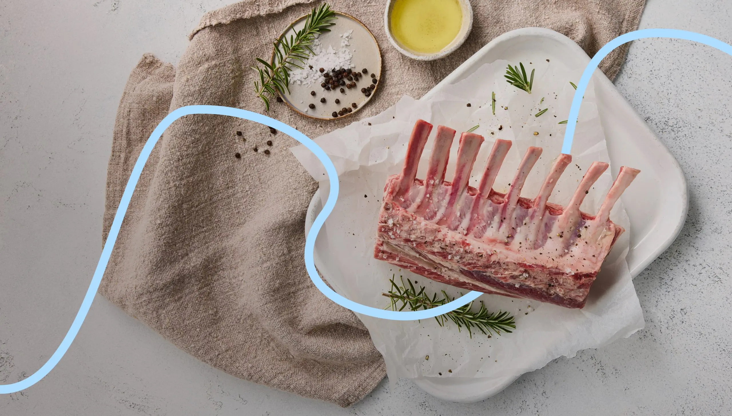 A fresh rack of lamb displayed on a white plate with parchment paper, surrounded by a linen cloth, olive oil, salt, black pepper, and fresh rosemary for seasoning.