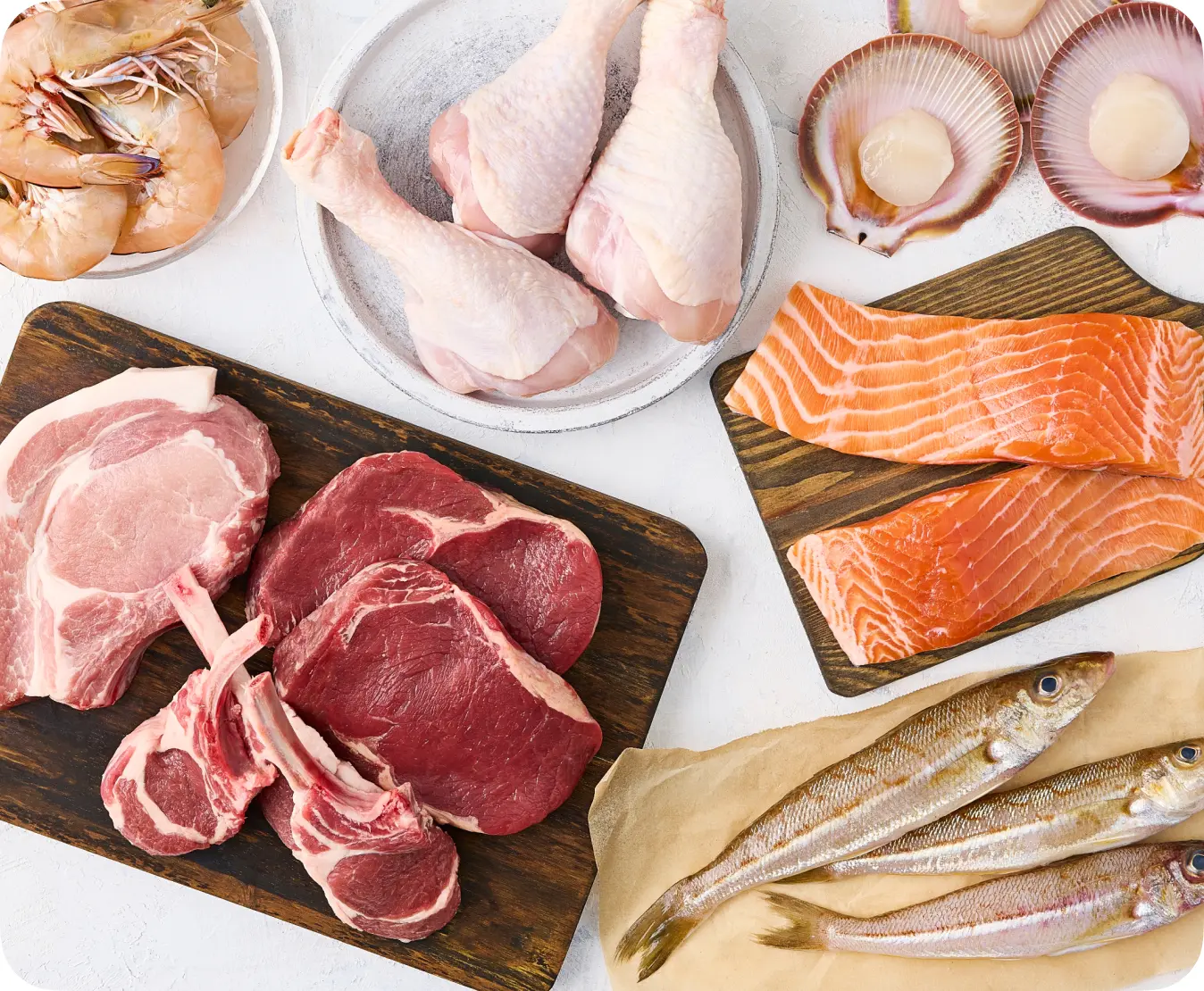 Assorted raw meats and seafood arranged on a white background, featuring chicken drumsticks, pork chops, beef cuts, salmon fillets, shrimp, and scallops, ideal for meal preparation and healthy cooking.
