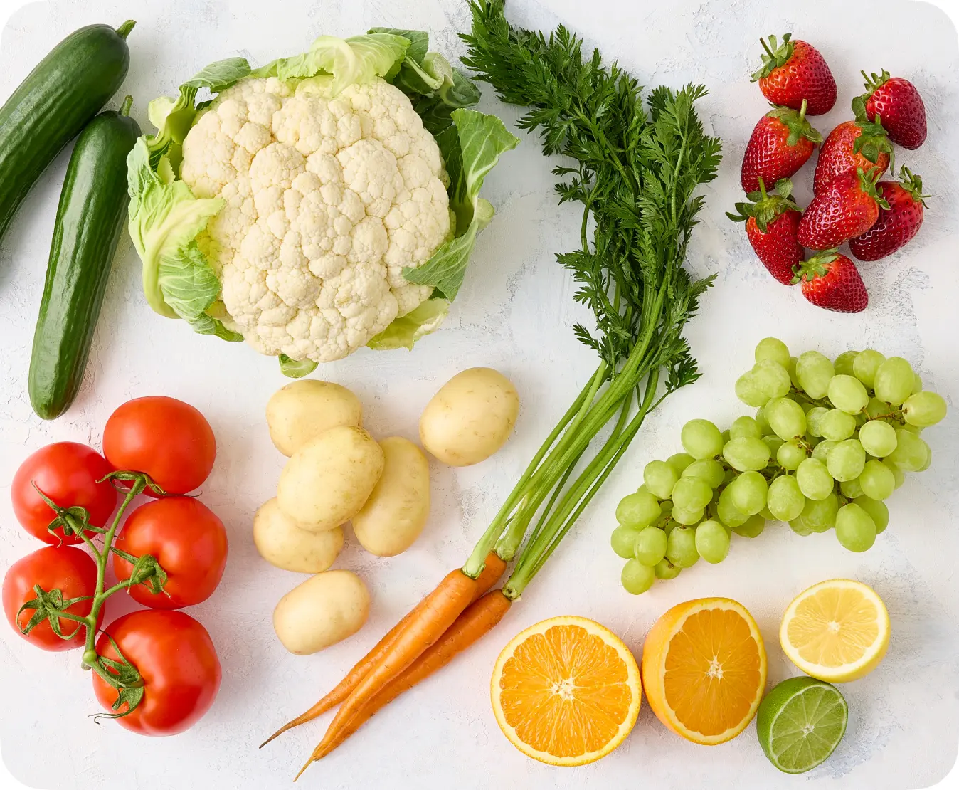 Fresh assortment of vegetables and fruits including cucumbers, cauliflower, tomatoes, potatoes, carrots, strawberries, green grapes, and citrus fruits like oranges, lemons, and limes, arranged on a light textured background, ideal for healthy eating and cooking inspiration.