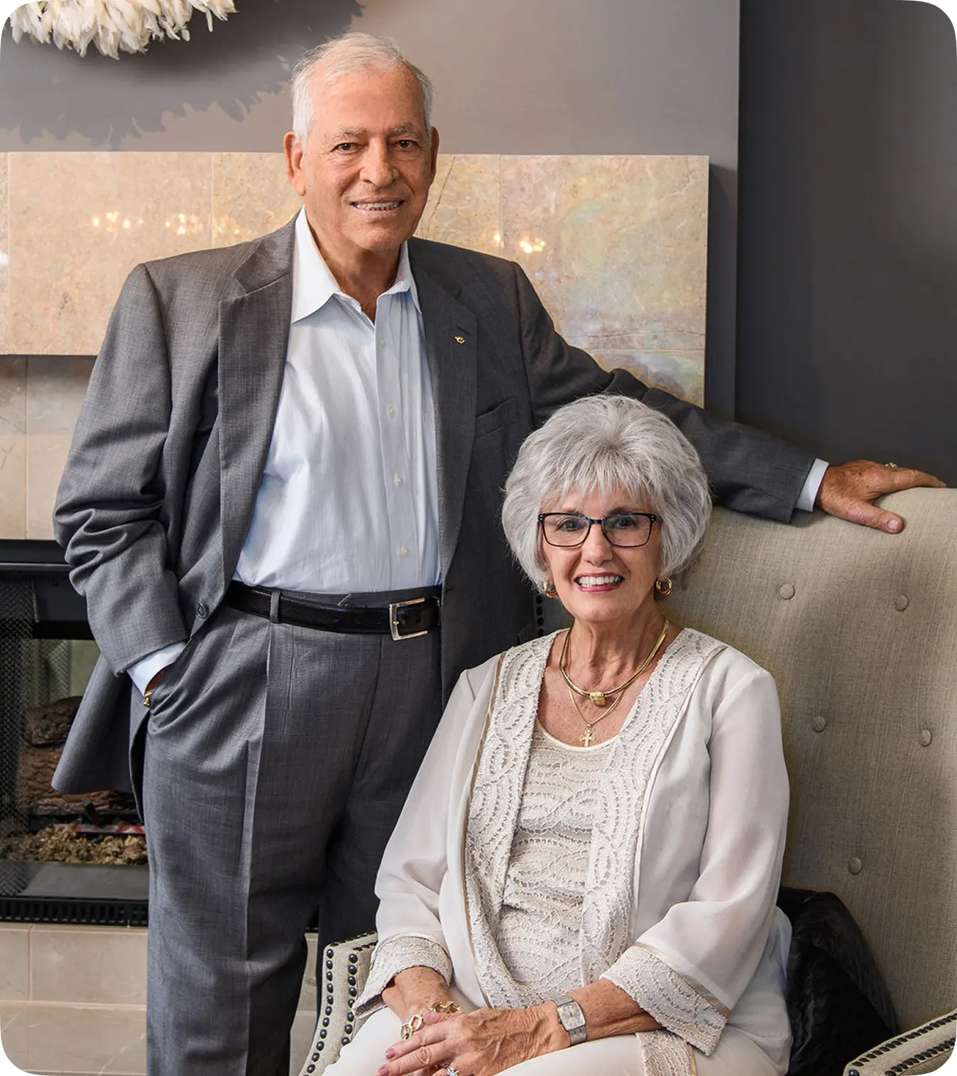 Elegant older couple posing indoors near a fireplace, smiling for the camera.