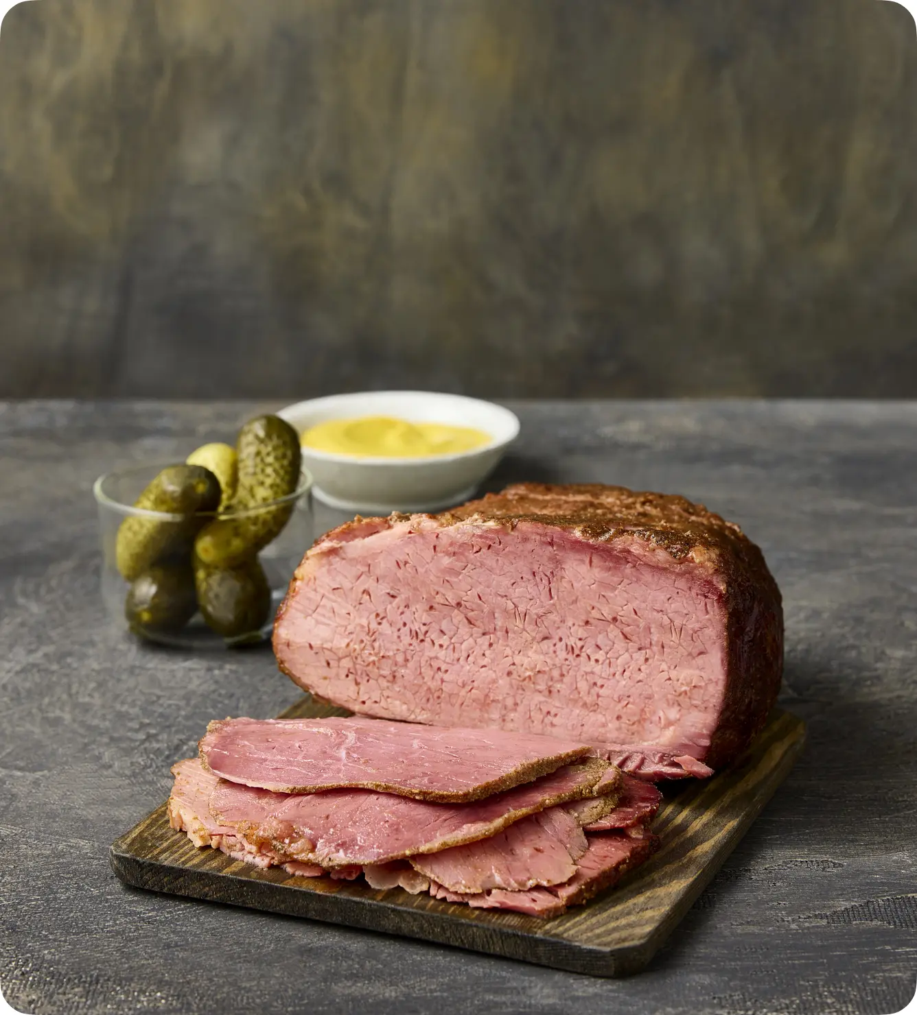Sliced corned beef on a wooden cutting board, accompanied by pickles and mustard in small bowls, set against a textured gray background.