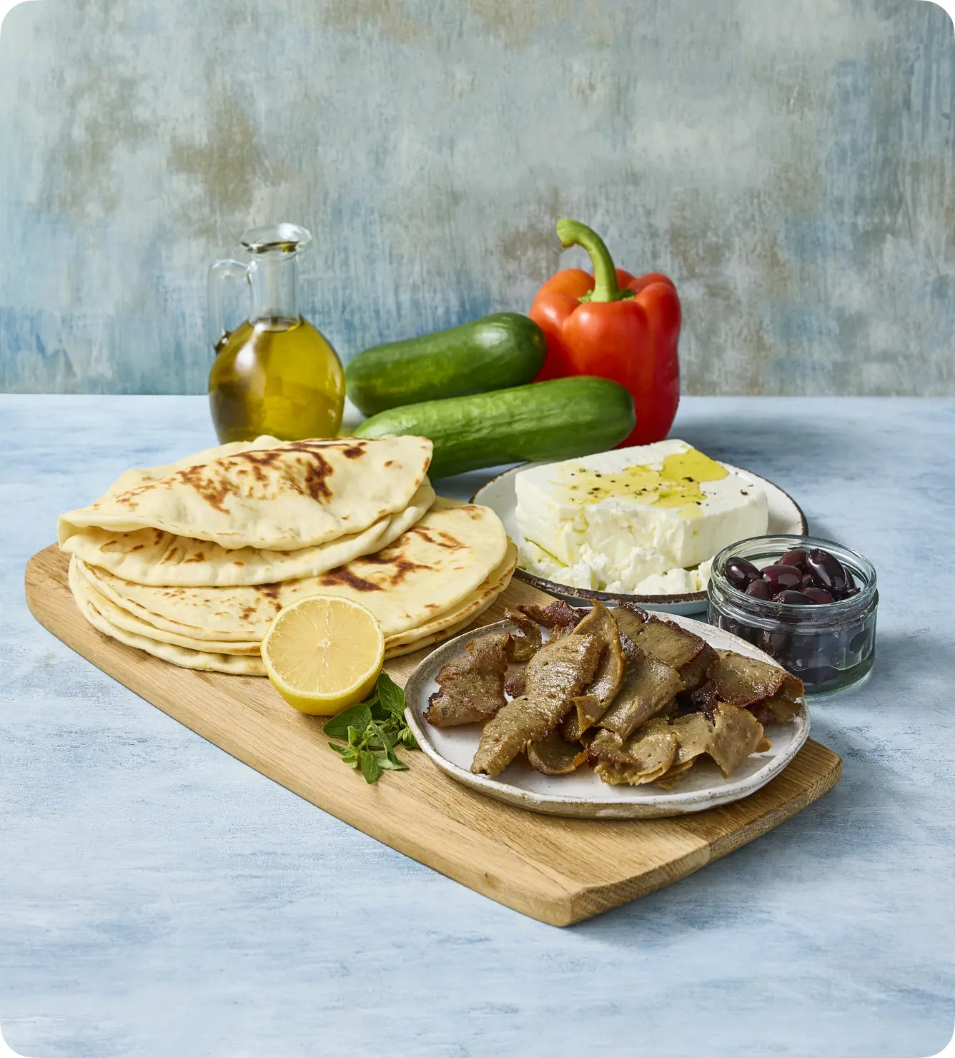 A wooden platter featuring grilled flatbreads, a plate of sliced meat, fresh cucumber, a red bell pepper, a block of feta cheese drizzled with olive oil, and a jar of black olives, with a lemon and fresh herbs for garnish.