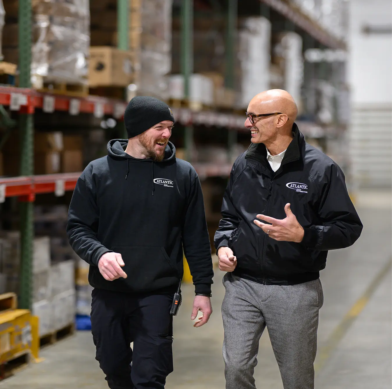 Two employees smiling and engaging in a conversation while walking through a warehouse filled with shelves of products, showcasing a positive workplace environment and teamwork.