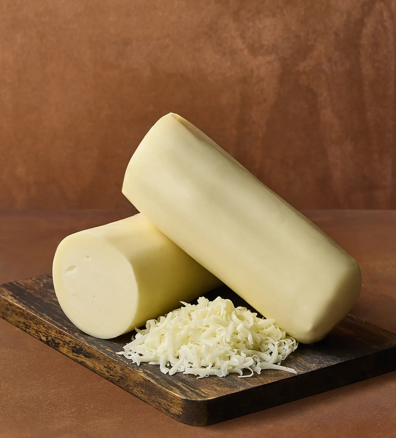 Fresh mozzarella cheese logs and shredded mozzarella on a wooden cutting board against a warm brown background, perfect for pizza and pasta dishes.