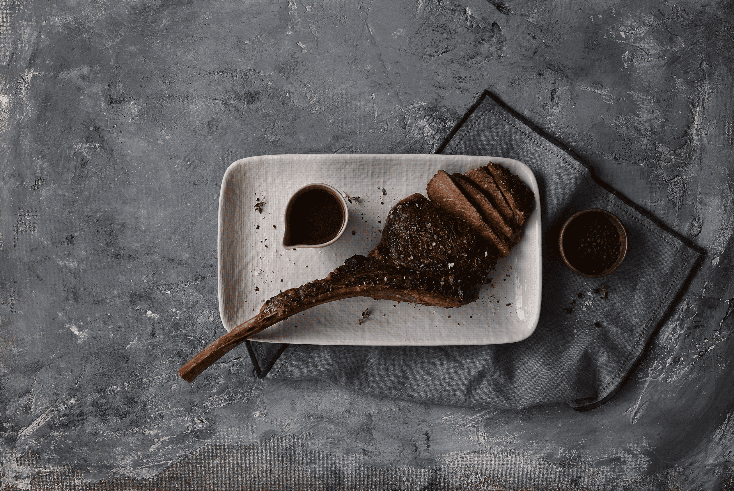 A beautifully presented tomahawk steak on a textured white plate, accompanied by a small jug of sauce and a dish of black pepper, set against a rustic gray background.