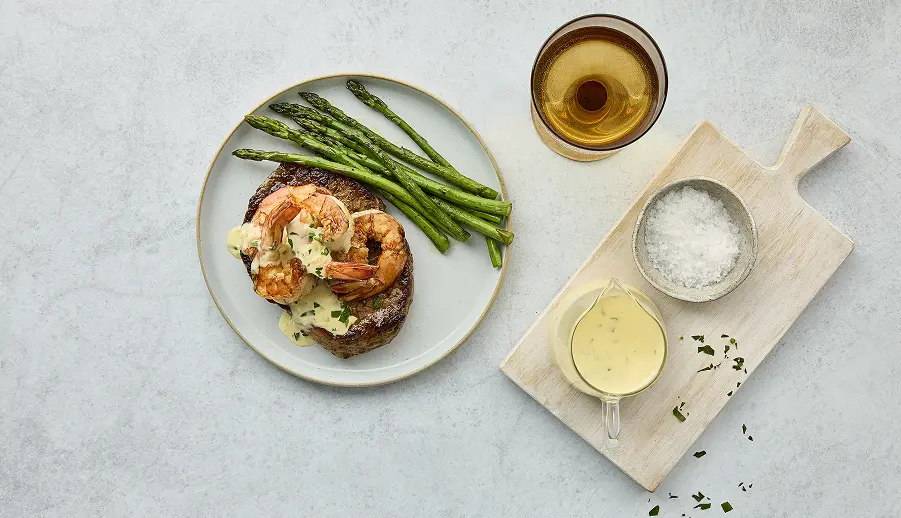 Grilled shrimp and steak topped with creamy sauce, served with asparagus on a white plate, accompanied by a glass of white wine, a bowl of sea salt, and a small dish of sauce on a wooden cutting board.