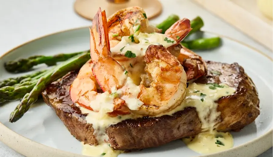 Grilled steak topped with succulent shrimp in a creamy sauce, garnished with fresh herbs, served alongside vibrant asparagus on a white plate. Perfect for a gourmet dinner or special occasion.