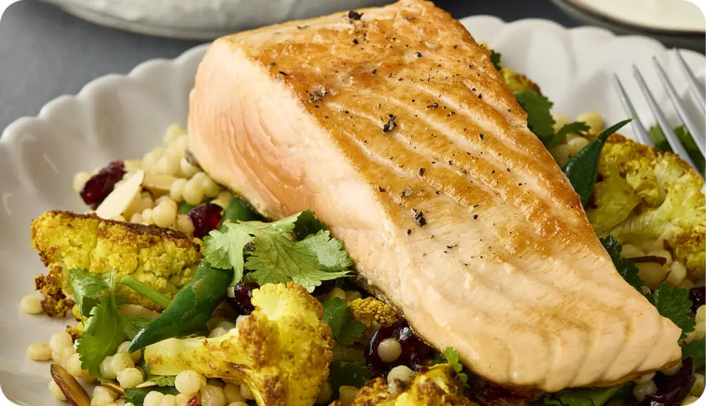 Grilled salmon fillet served with couscous, roasted cauliflower, green beans, and fresh herbs on a white plate