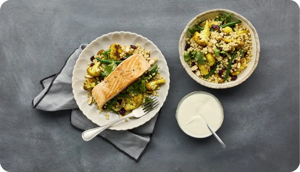 Top view of grilled salmon with couscous, roasted vegetables, and yogurt sauce served on a table with side dishes