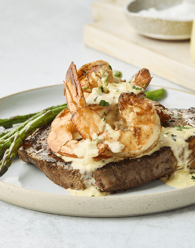 Grilled steak topped with sautéed shrimp and creamy sauce, served with asparagus on a white plate.