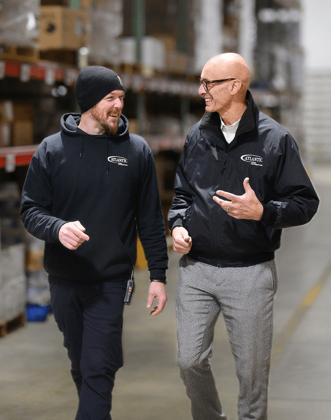 Two men walking and talking in a warehouse, wearing branded jackets, illustrating teamwork and collaboration in a business environment. The background features shelves stocked with goods, highlighting a productive workspace.