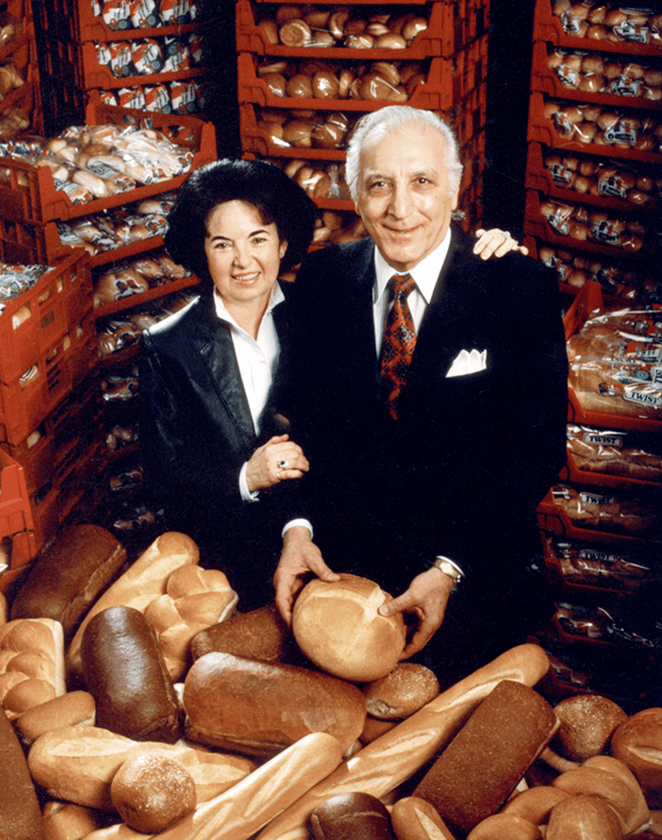A couple stands proudly among a variety of freshly baked bread loaves and rolls, with stacks of bread crates in the background, showcasing their successful bakery business.