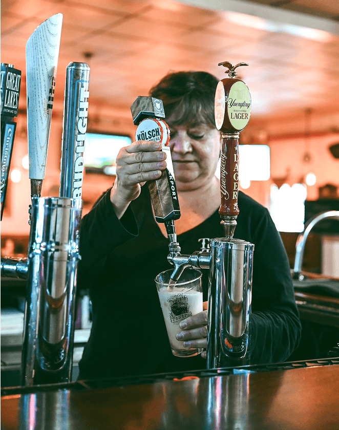 Bartender pouring a Kölsch beer into a glass from a tap at a bar, showcasing various beer taps in the background, with a focus on the beer being served.