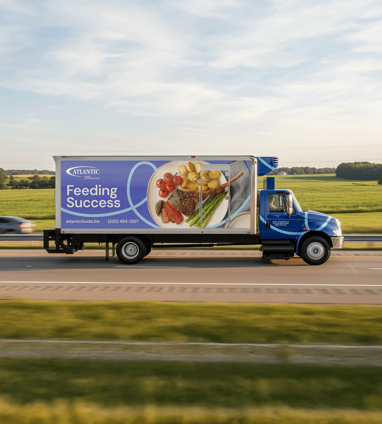 Delivery truck from Atlantic Foods driving on a highway, featuring a colorful advertisement showcasing a plated meal with steak, potatoes, cherry tomatoes, and asparagus. The tagline "Feeding Success" is prominently displayed along with contact information and website. Lush green fields in the background.